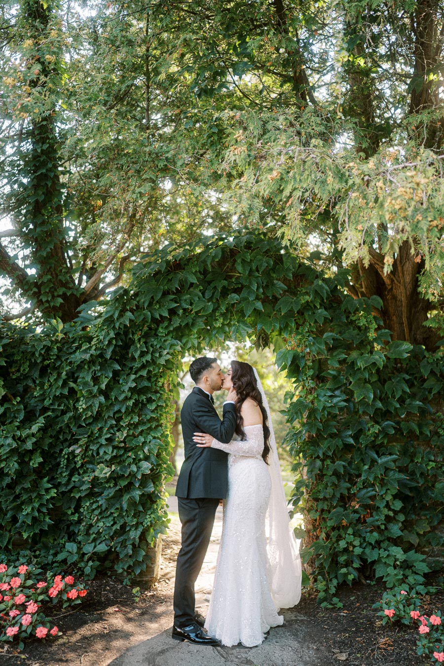 A bride and groom share a kiss under a lush, green ivy archway in a garden setting, surrounded by blooming flowers and