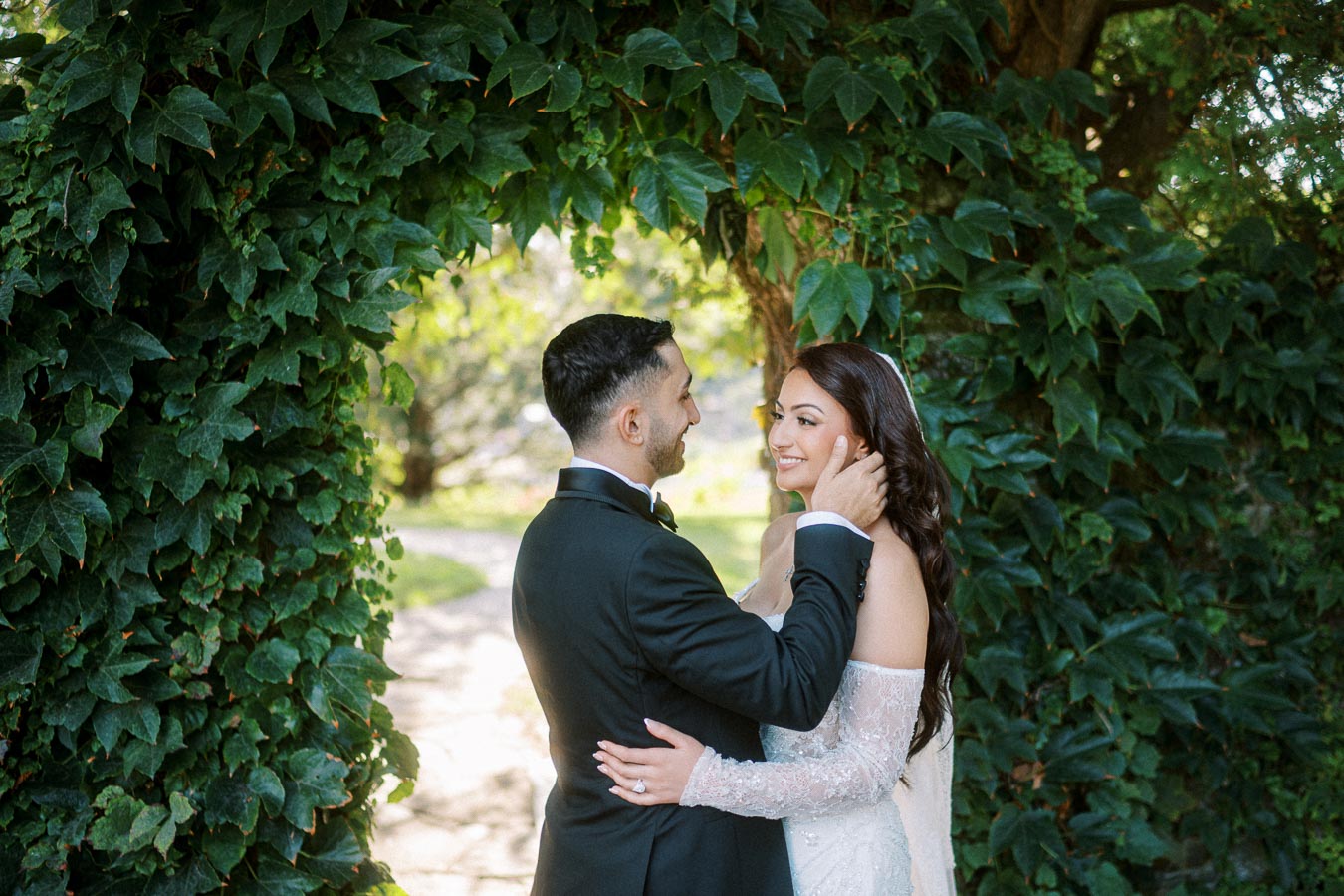 Bride and groom smiling at each other under a lush green archway, celebrating their wedding day in a picturesque garden