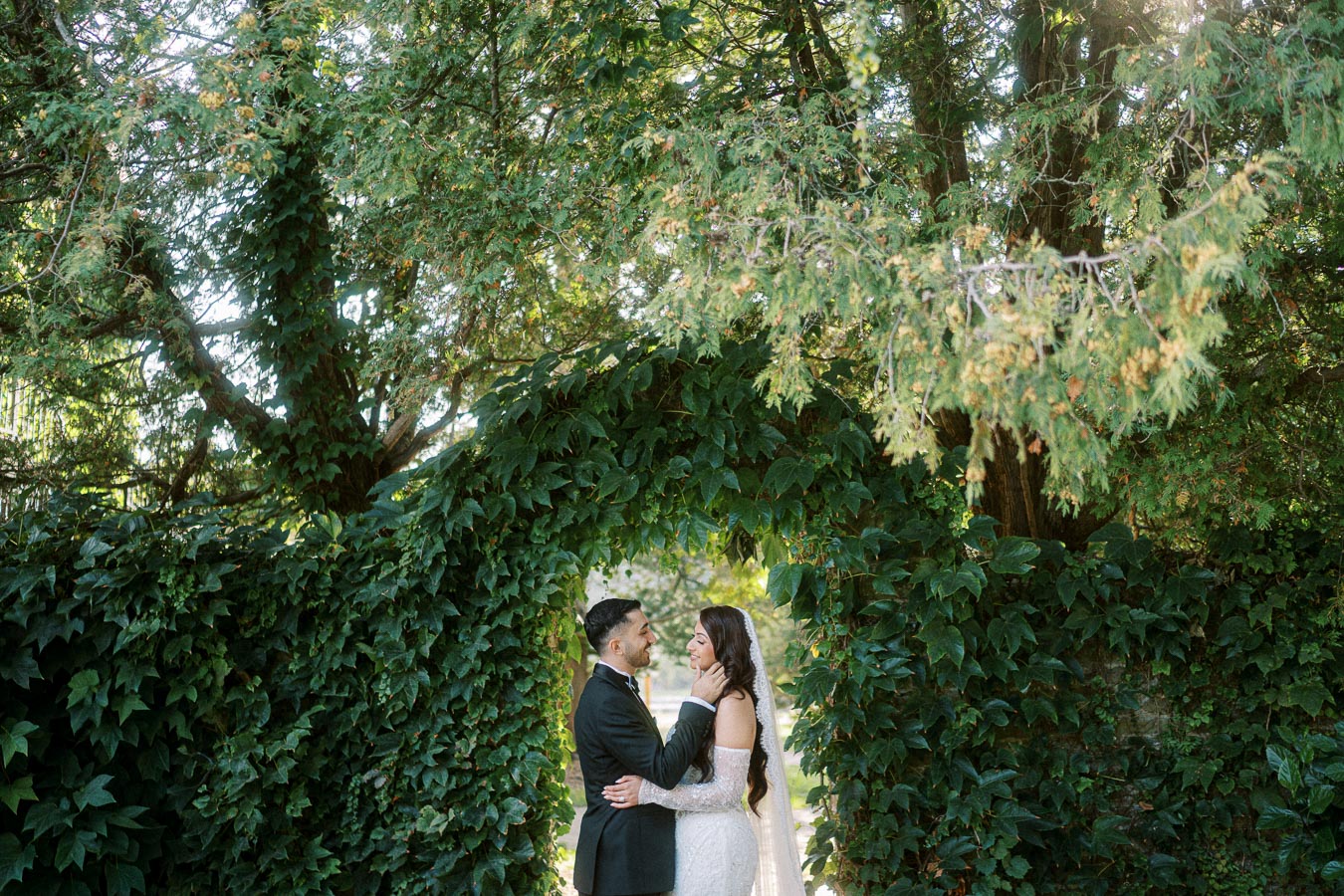 Bride and groom gazing at each other under a lush green archway in a sunlit garden.