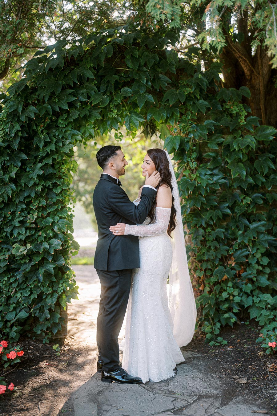 A bride and groom smiling at each other under a lush green ivy archway in a garden, captured in a candid wedding moment.