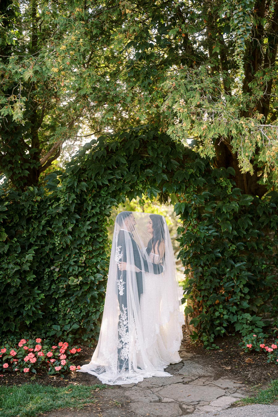 A bride and groom share an intimate moment under a lush green archway, wrapped in a delicate lace veil, surrounded by