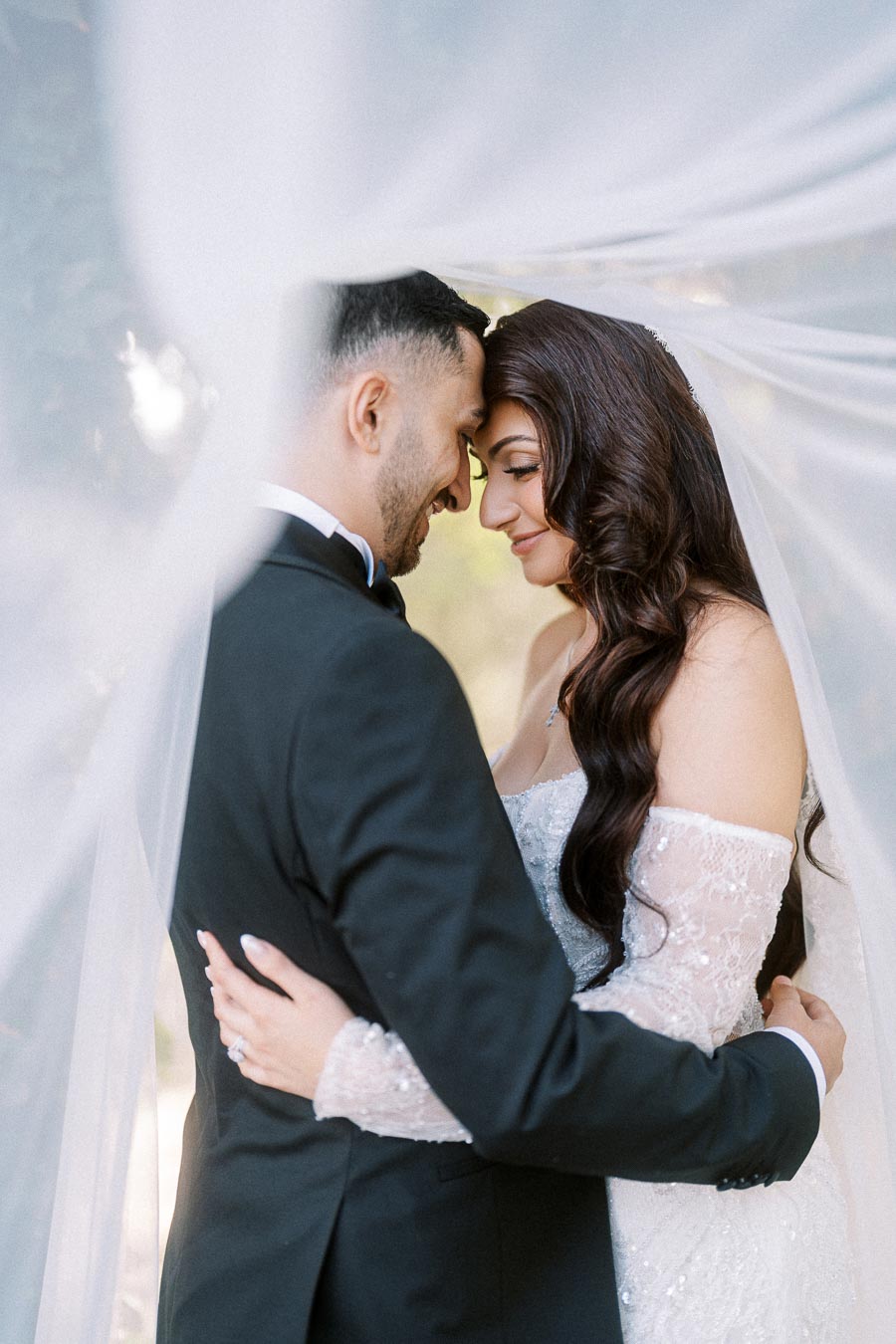 A newlywed couple embracing tenderly under a flowing white veil, with the groom in a classic black suit and the bride in an