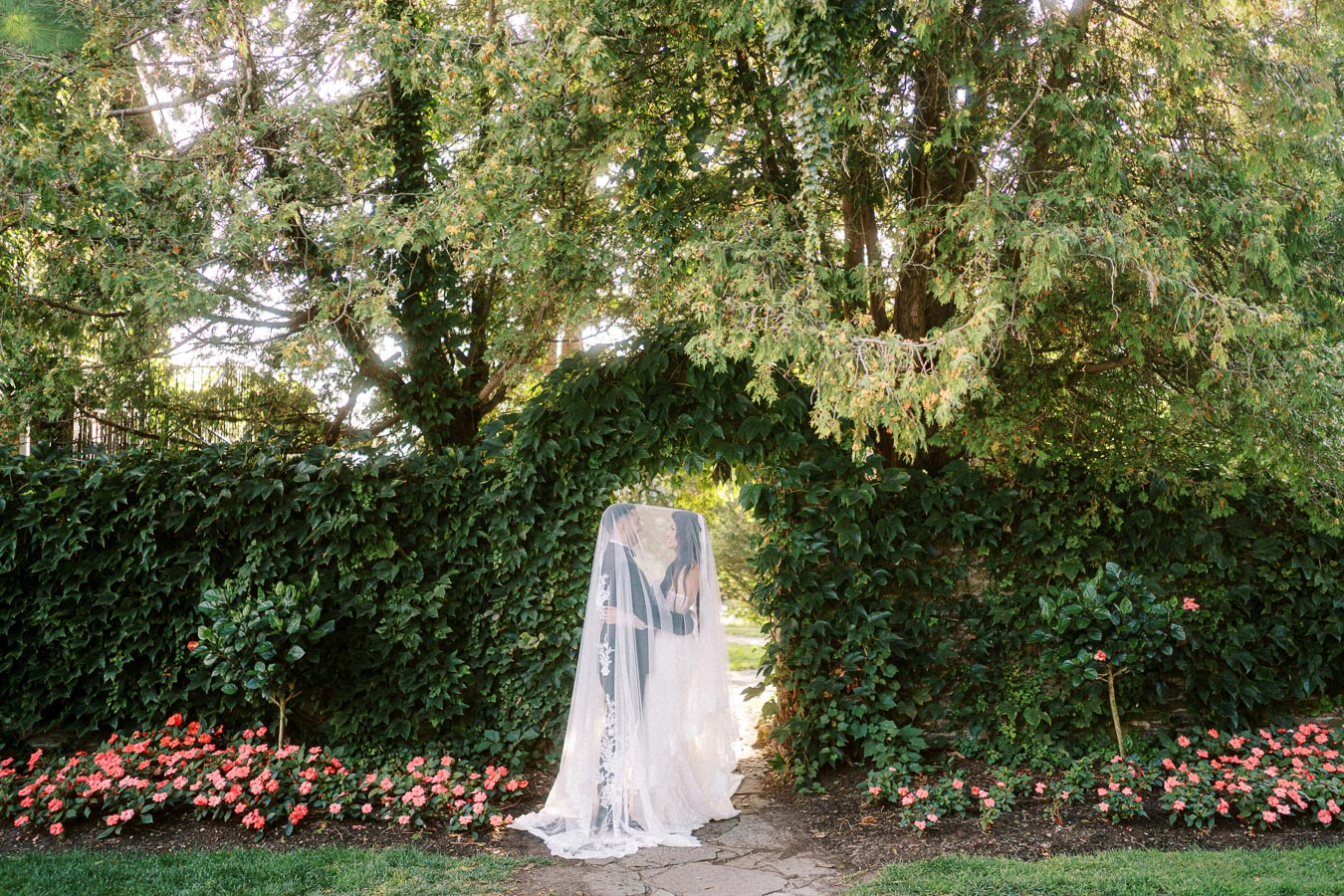 A bride and groom share a romantic moment beneath a veil in a lush garden setting with vibrant pink flowers and verdant
