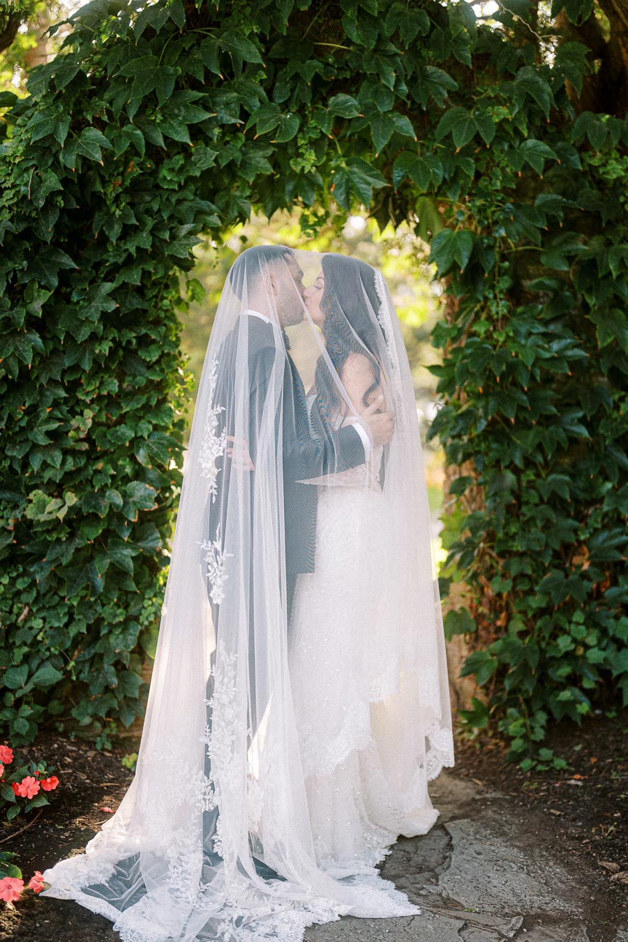 A bride and groom share a kiss under a veil in a lush garden setting, surrounded by vibrant green ivy and blooming flowers,