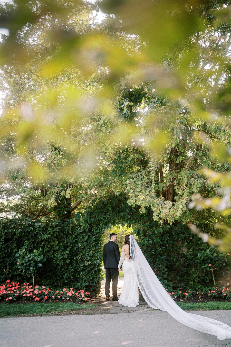 A bride in a flowing white wedding dress and long veil stands hand in hand with a groom in a black suit, posing in front of