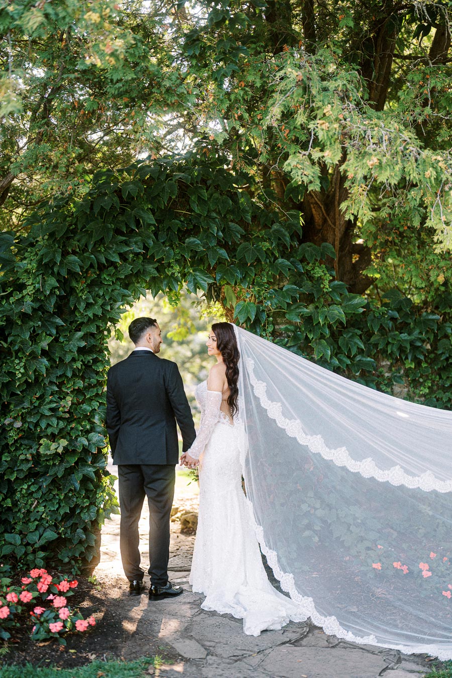 A bride in a long lace wedding gown and veil holds hands with a groom in a black suit, standing beneath a lush green archway