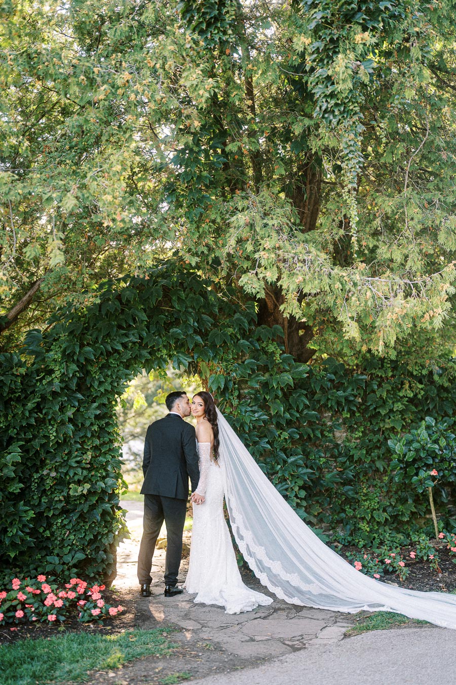 A bride and groom stand hand in hand under a lush green archway, surrounded by vibrant foliage. The bride wears a stunning