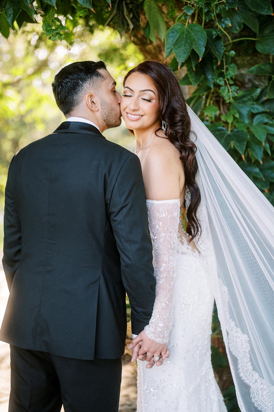 Romantic outdoor wedding photo of a groom kissing the bride's cheek, standing hand-in-hand under a leafy tree, with the