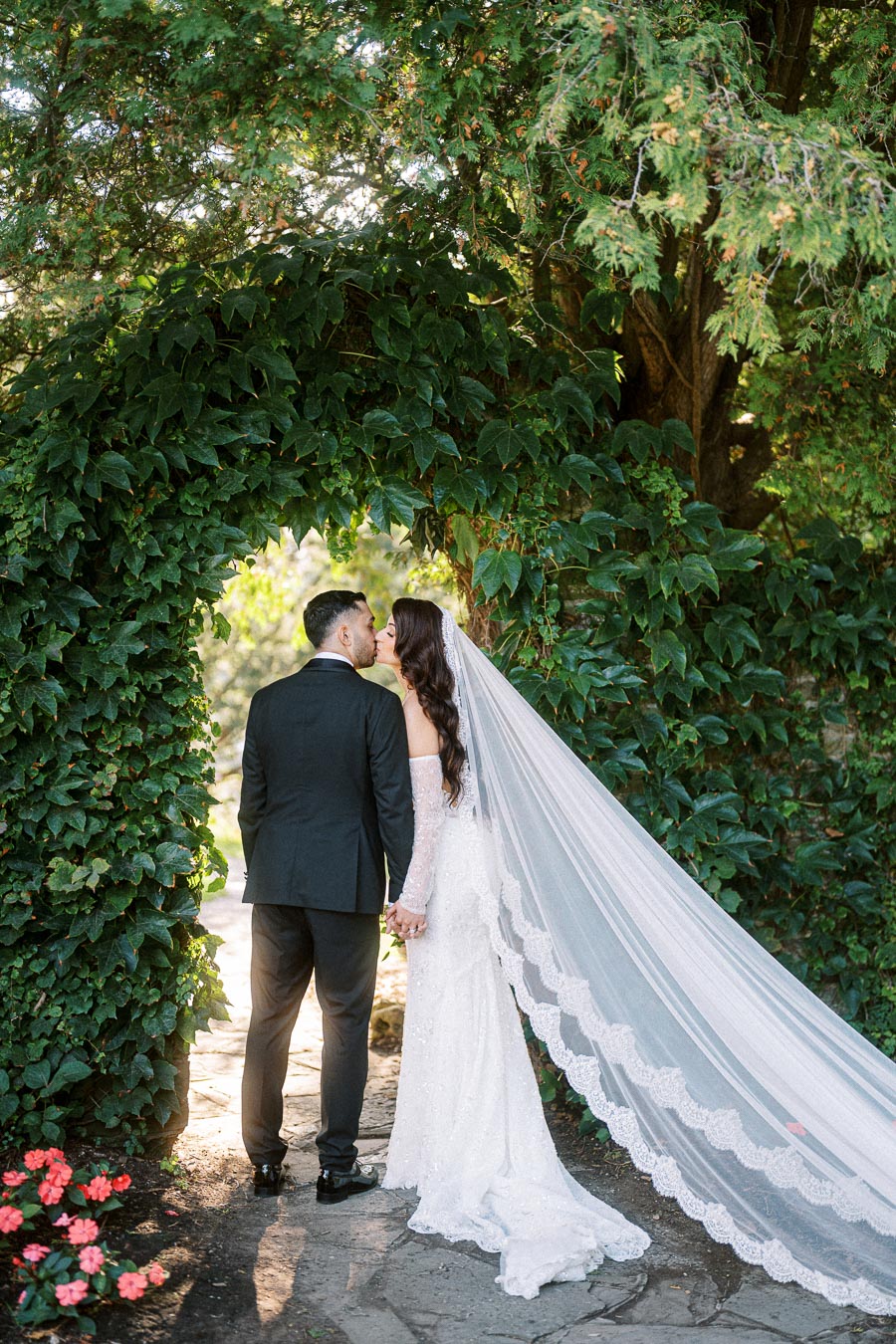 Bride and groom kissing under a lush greenery archway at an outdoor garden wedding, with the bride in a flowing white gown
