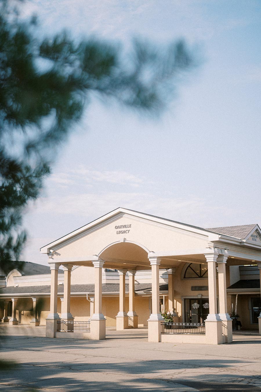 Front view of a beige building with columns labeled 'Oakville Legacy' under a clear blue sky, framed by blurred