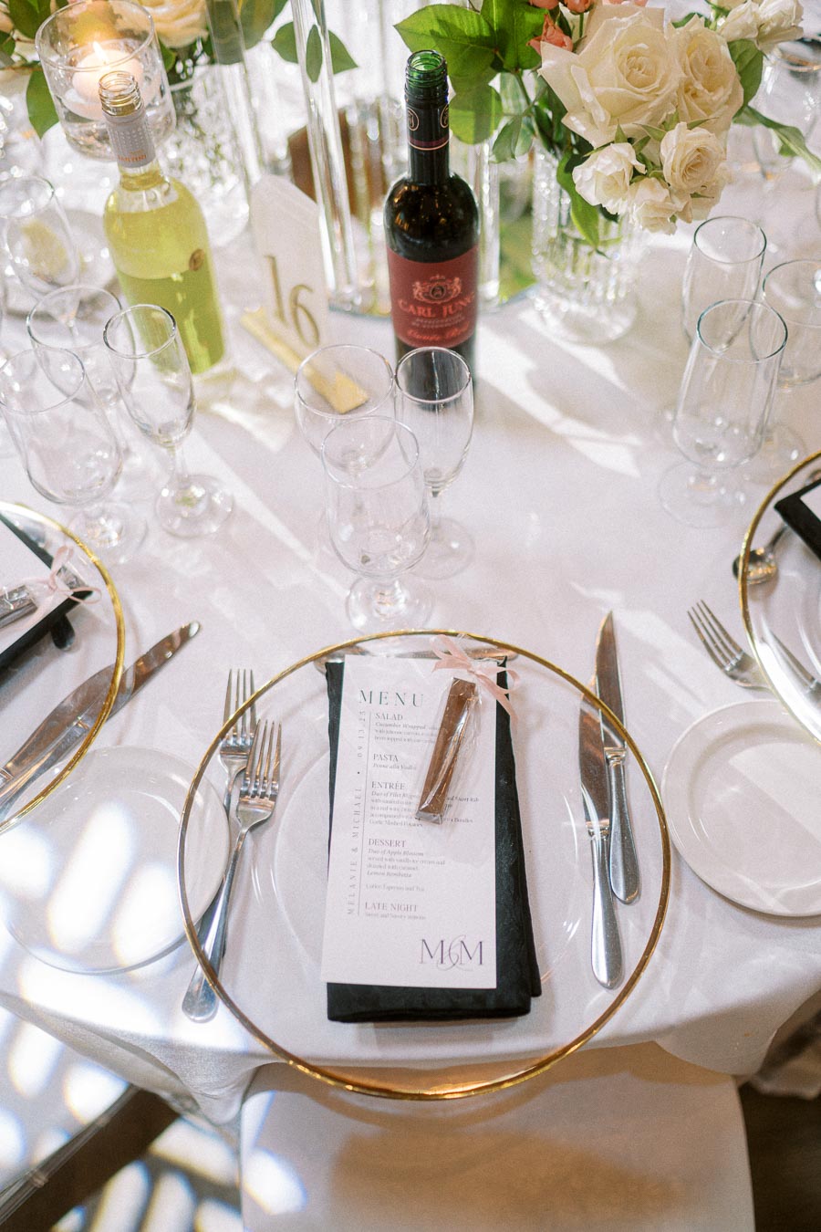 Elegant wedding table setting with wine bottles, flowers, crystal glassware, and a detailed menu on a white tablecloth.