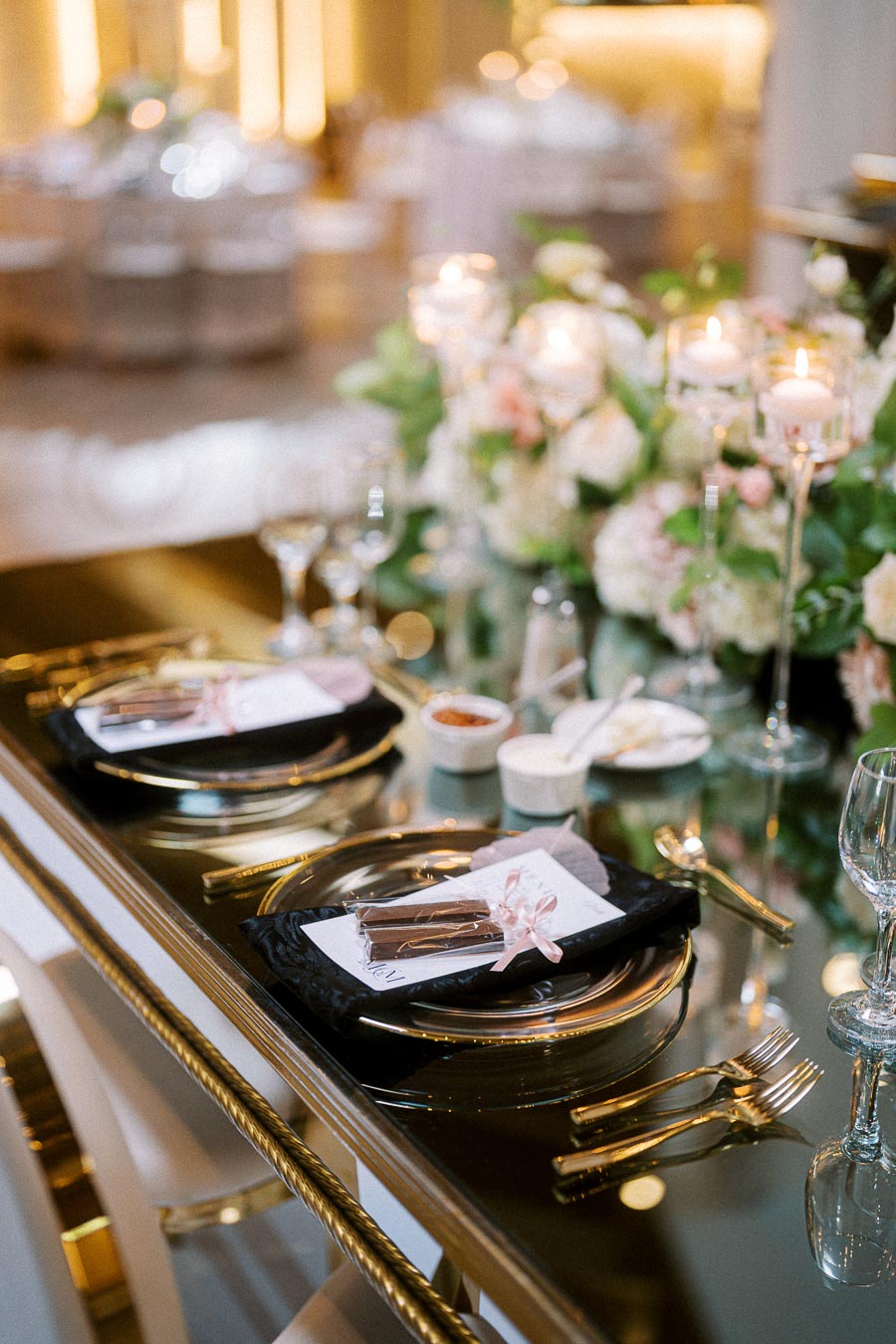 Elegant wedding table setting with glass plates, gold cutlery, and black napkins on a mirrored surface, adorned with candles