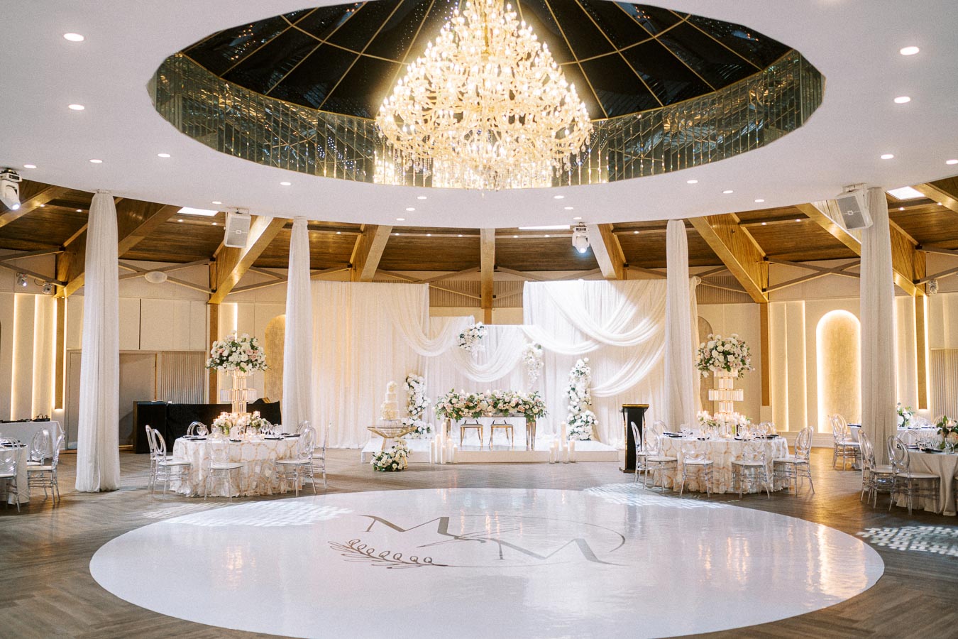 Elegant wedding reception venue featuring a grand chandelier, draped white curtains, and floral arrangements. The modern