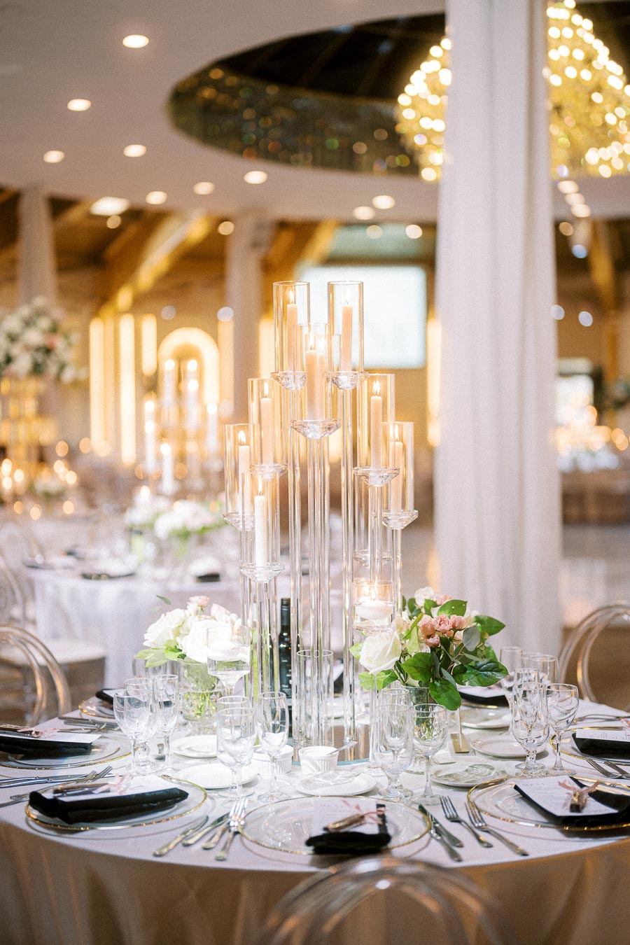 Elegant wedding reception table setting with tall glass candle holders, glowing candles, floral centerpieces, and neatly