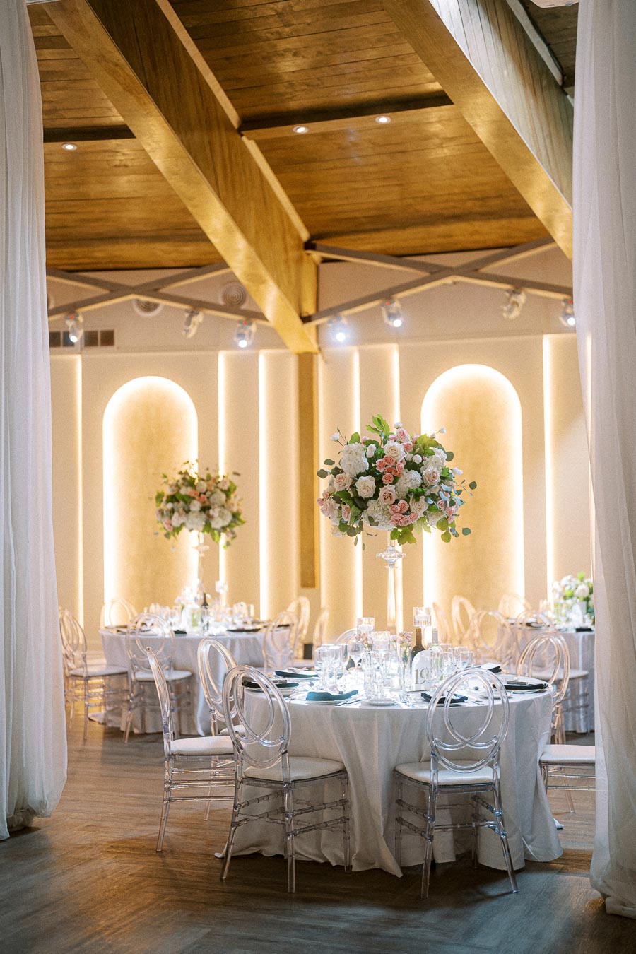 Elegant wedding reception venue with round tables, floral centerpieces, and transparent chairs, set in a warmly lit,