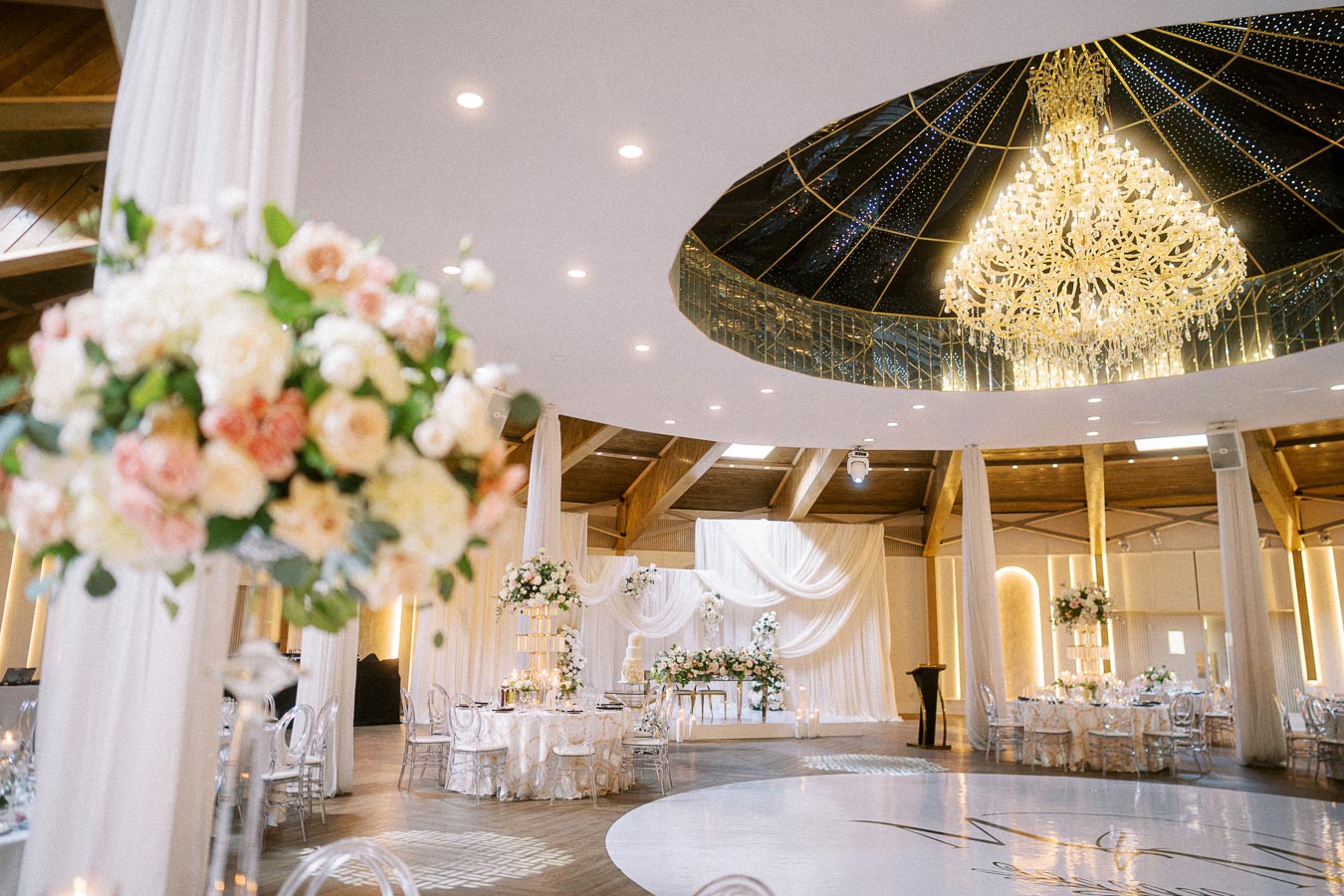 Elegant wedding venue with luxurious chandelier, round tables adorned with floral arrangements, and white draped curtains