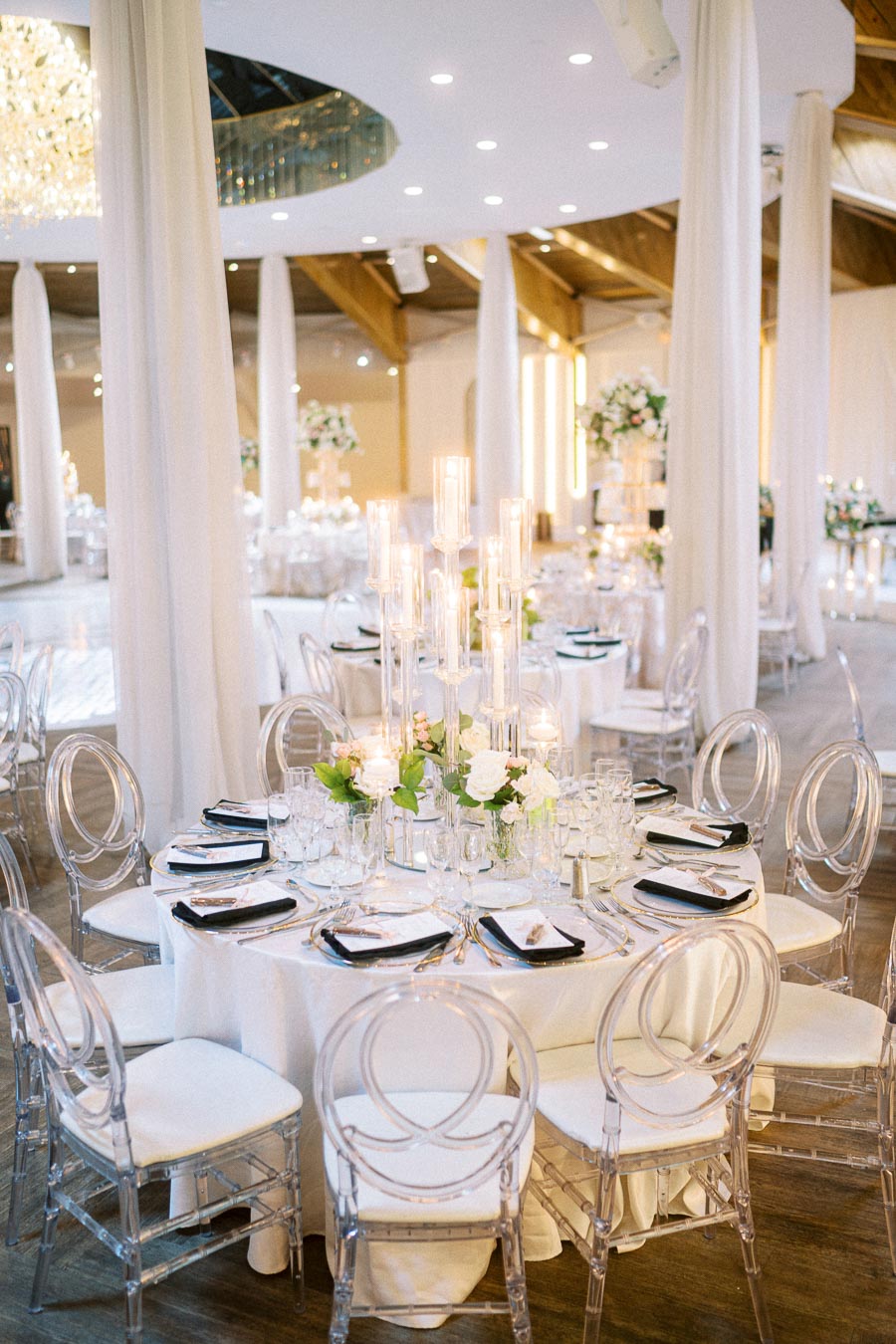 Elegant wedding reception table setting with clear chairs, tall glass candle holders, and floral centerpieces in a luxurious
