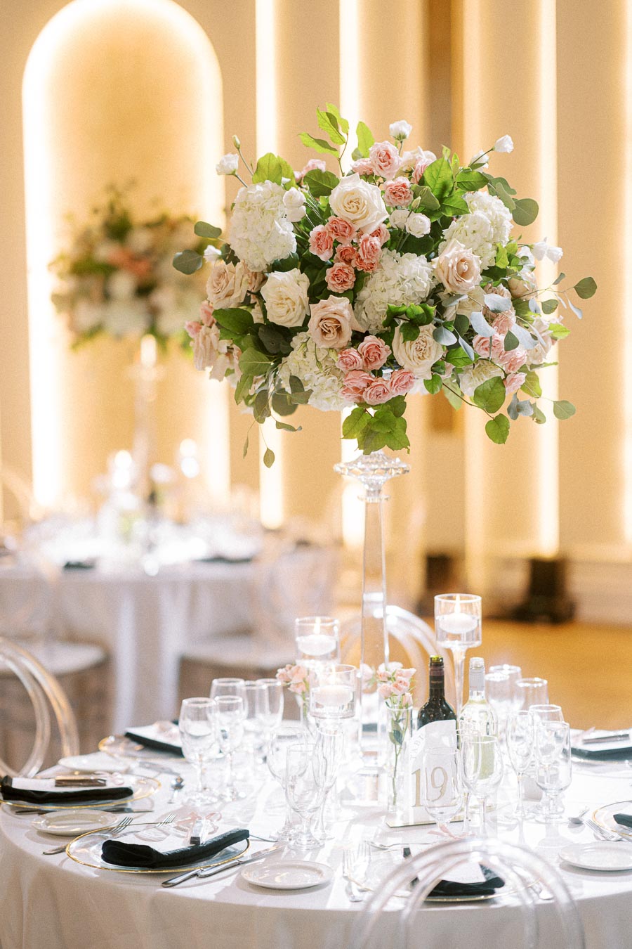 Elegant wedding reception table setting with tall floral centerpiece featuring white and pink roses, surrounded by