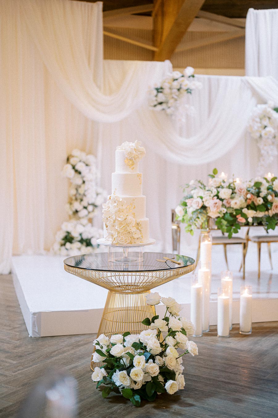 A luxurious four-tier white wedding cake adorned with elegant floral accents on a modern gold table; surrounded by white