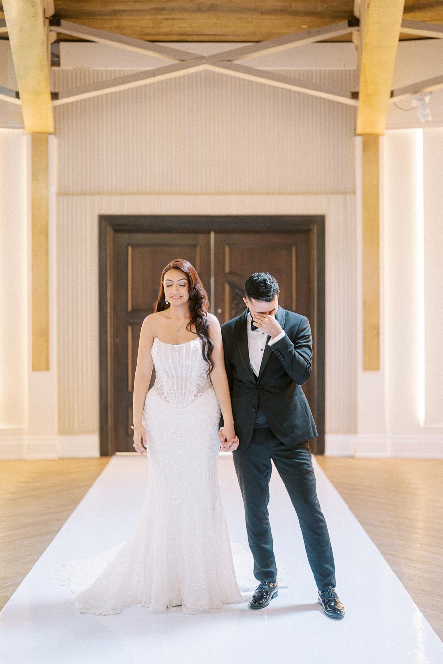 Elegant bride in a white gown holding hands with a groom in a tuxedo, posing in an elegantly decorated venue with wooden