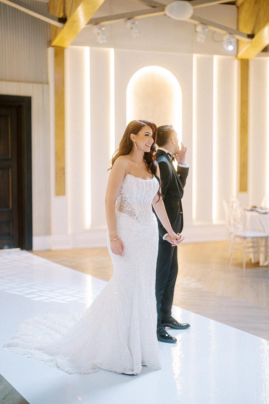 Bride in a lace wedding dress holding hands with a groom in a tuxedo, smiling during their elegant indoor wedding ceremony.