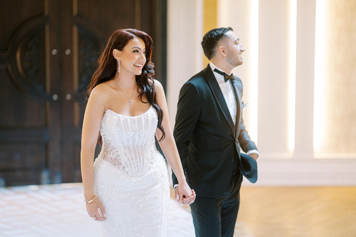 A bride in a white lace wedding dress and a groom in a black tuxedo joyfully holding hands in an elegant venue, highlighting