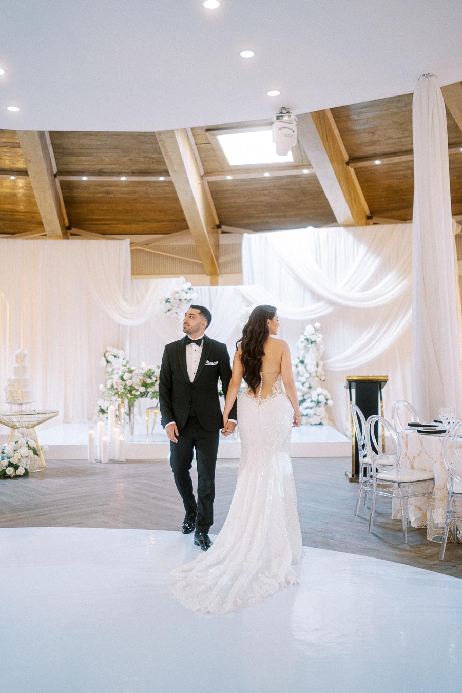 A bride and groom in elegant attire walking hand in hand at a beautifully decorated indoor wedding venue with draped fabric