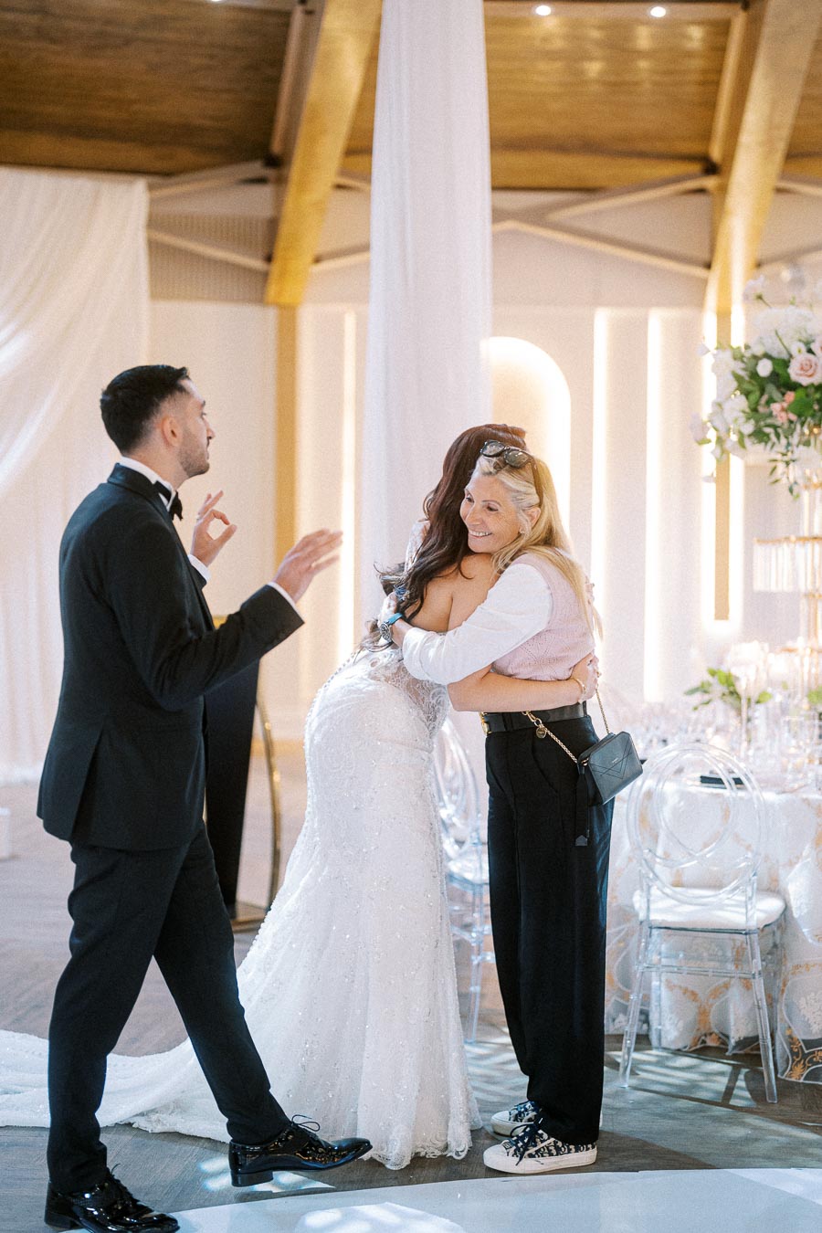 A groom in a black tuxedo gestures while a bride in a white wedding dress hugs a guest with long blonde hair at a wedding