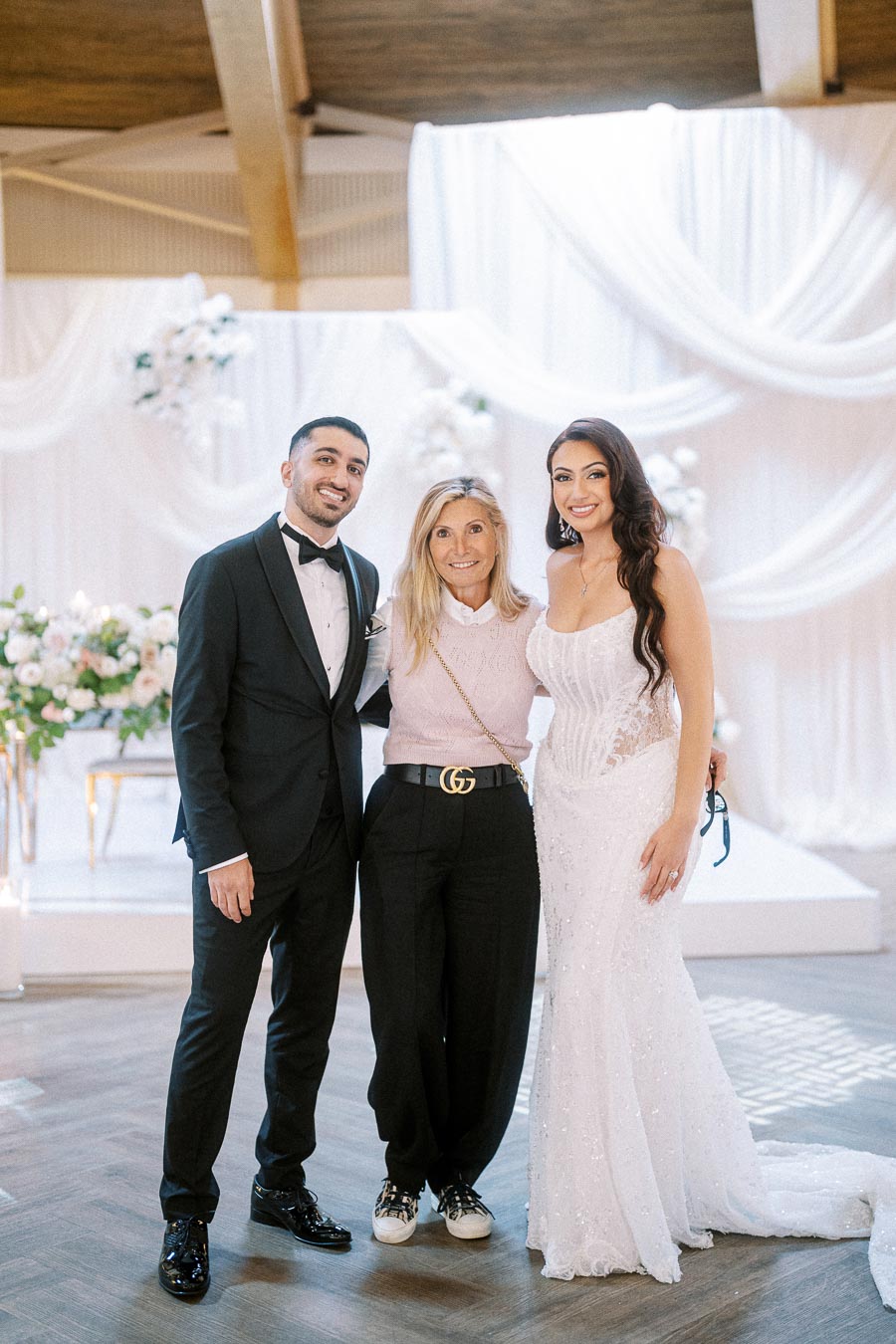 A bride in a white wedding dress stands alongside a groom in a black tuxedo and a woman in casual attire, posing together at