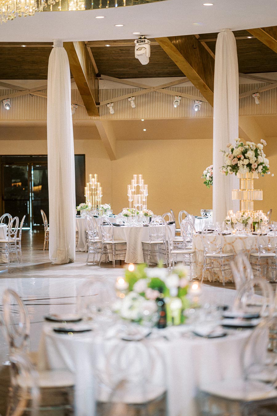 Elegant wedding reception hall with beautifully arranged round tables, transparent chairs, and tall floral centerpieces,