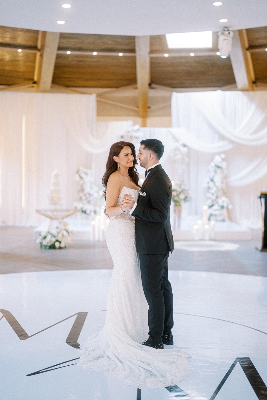 A newlywed couple shares their first dance in an elegant, modern wedding venue with white drapery and floral arrangements.