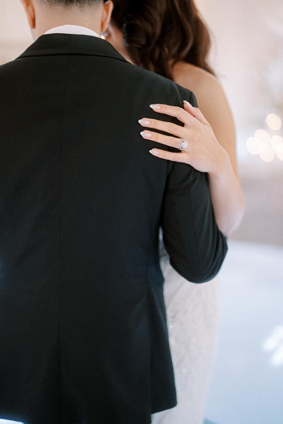 A bride and groom embrace in a tender moment, with the bride's hand resting on the groom's back, showcasing a sparkling