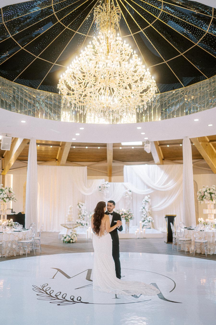 A couple shares their first dance under a luxurious chandelier in an elegantly decorated wedding venue with floral