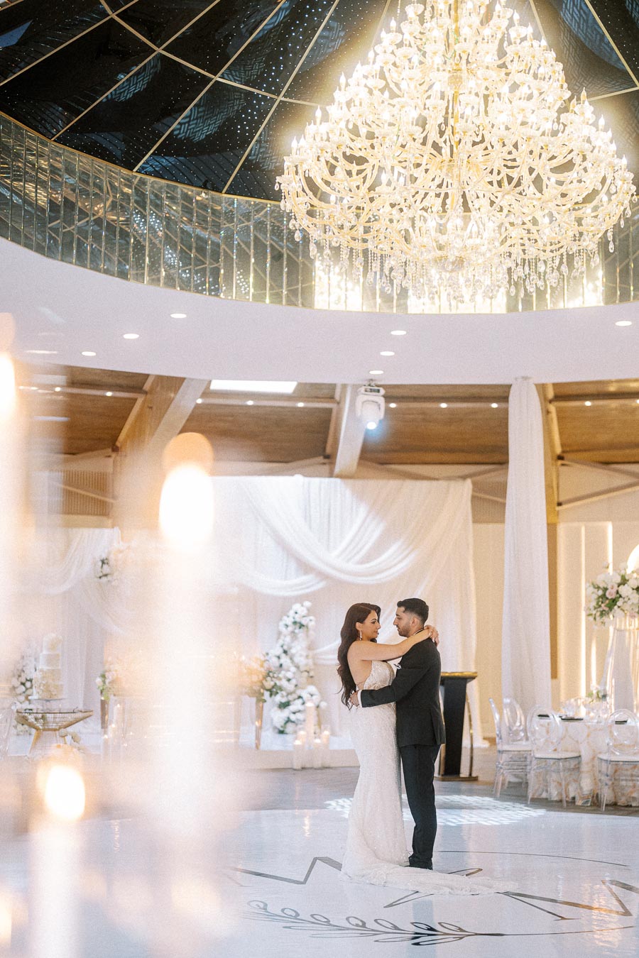 A newlywed couple dances closely under a magnificent chandelier in an elegantly decorated reception hall, featuring white