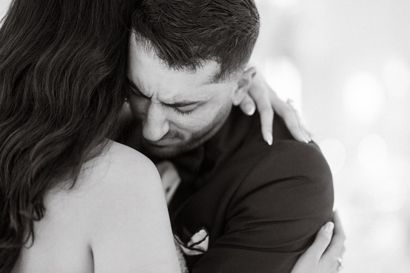 A black and white image of a couple sharing an emotional embrace, featuring a man in a suit with eyes closed and a woman