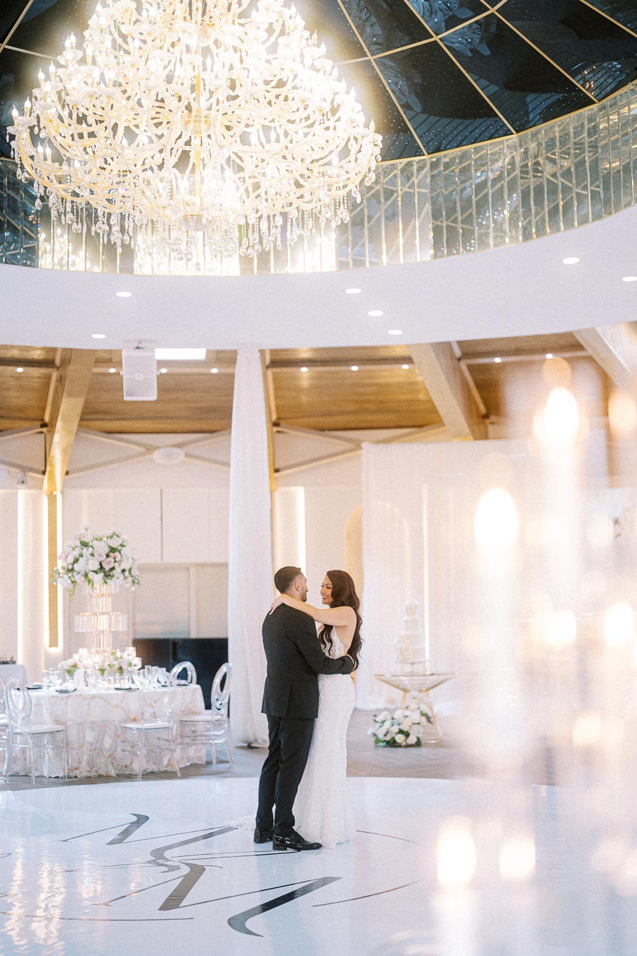 A couple embraces on an elegant dance floor beneath a sparkling chandelier at a beautifully decorated wedding venue.