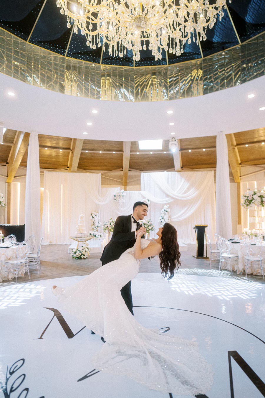 Elegant wedding couple dancing under a luxurious chandelier in a beautifully decorated venue with draped curtains and floral