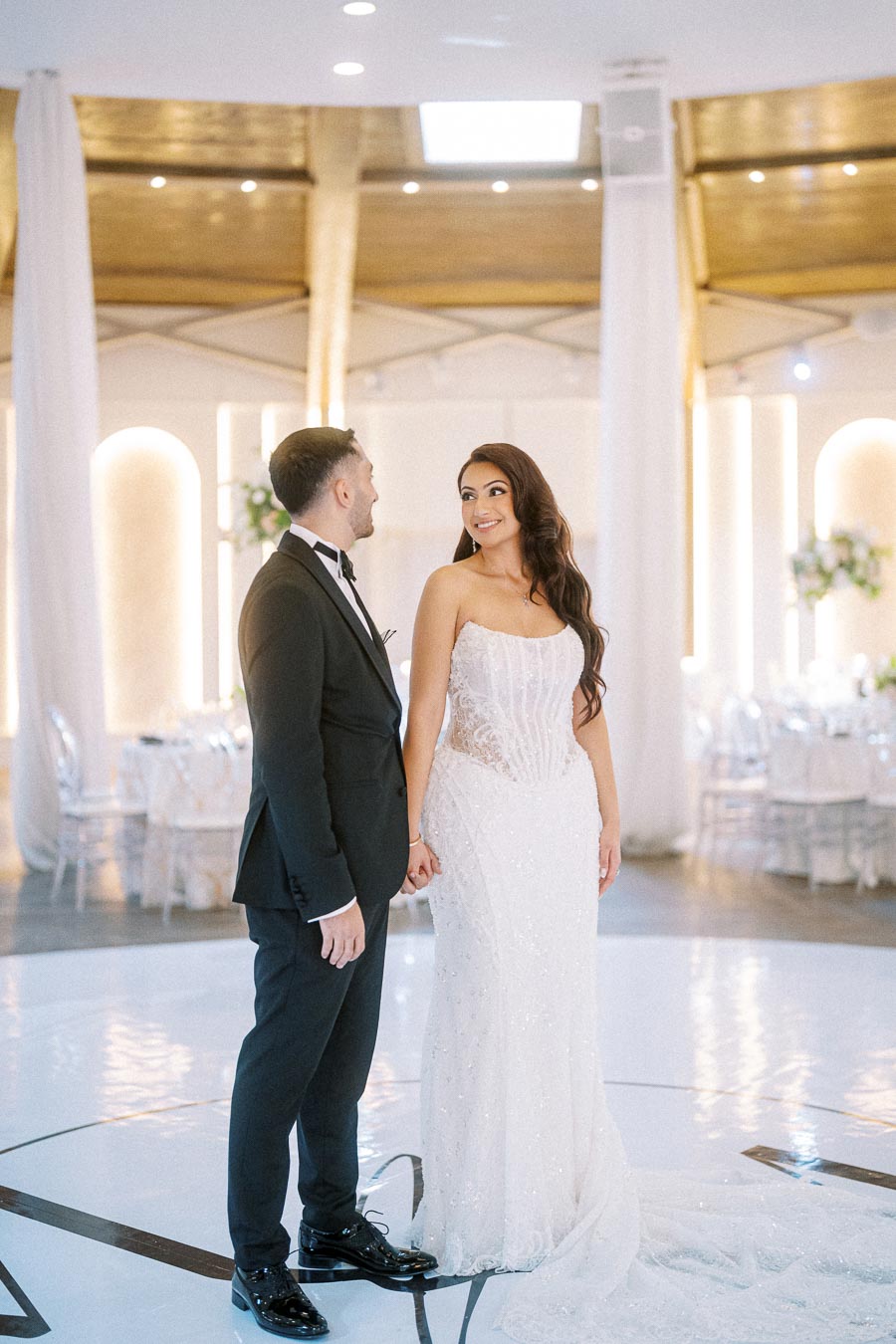 Elegant bride in a strapless white gown and groom in a black tuxedo holding hands, standing in a beautifully decorated