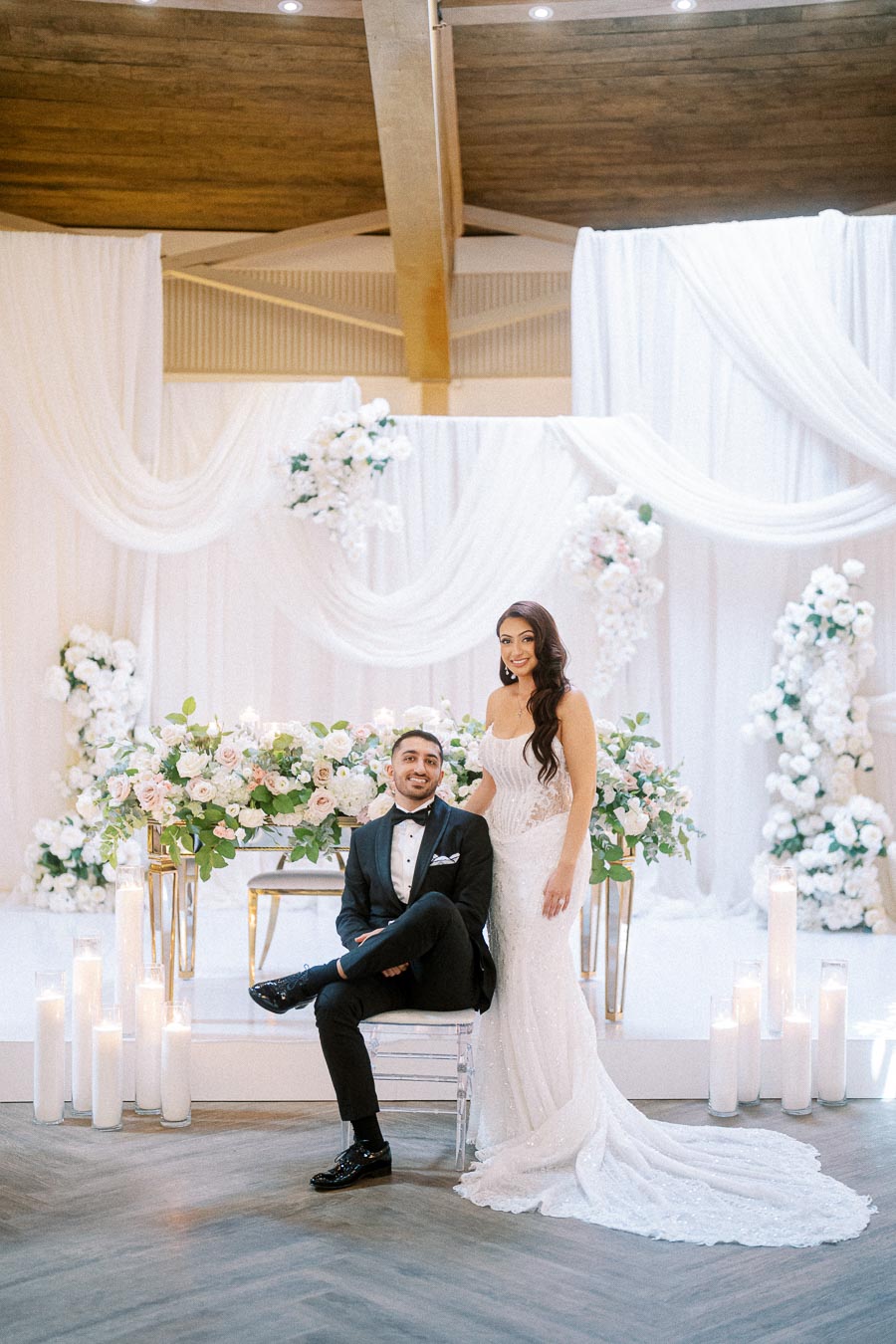 A bride in a white lace gown stands beside a seated groom in a black tuxedo, in front of an elegant floral and draped