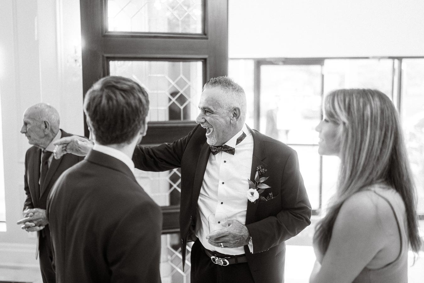 A joyful man in a tuxedo interacts with guests at a formal gathering, holding a drink, with smiling participants in an