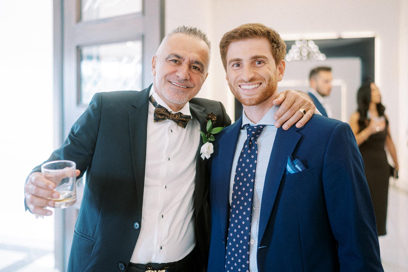 Two men in suits smiling warmly at a celebration event; one holds a drink and wears a black bow tie and white boutonniere,