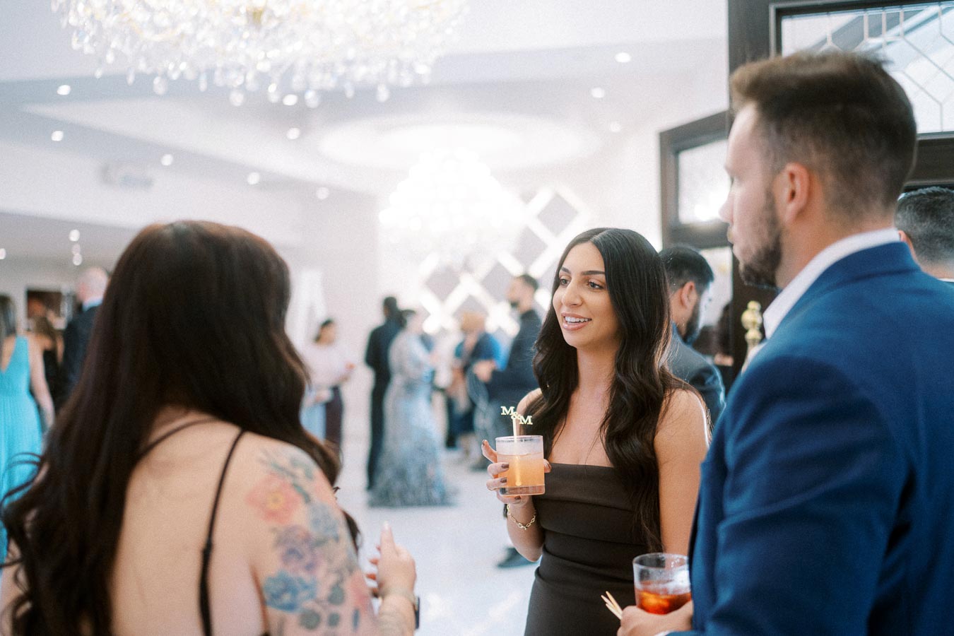 A group of elegantly dressed people socializing at a formal event, with a woman holding a cocktail and smiling, set against