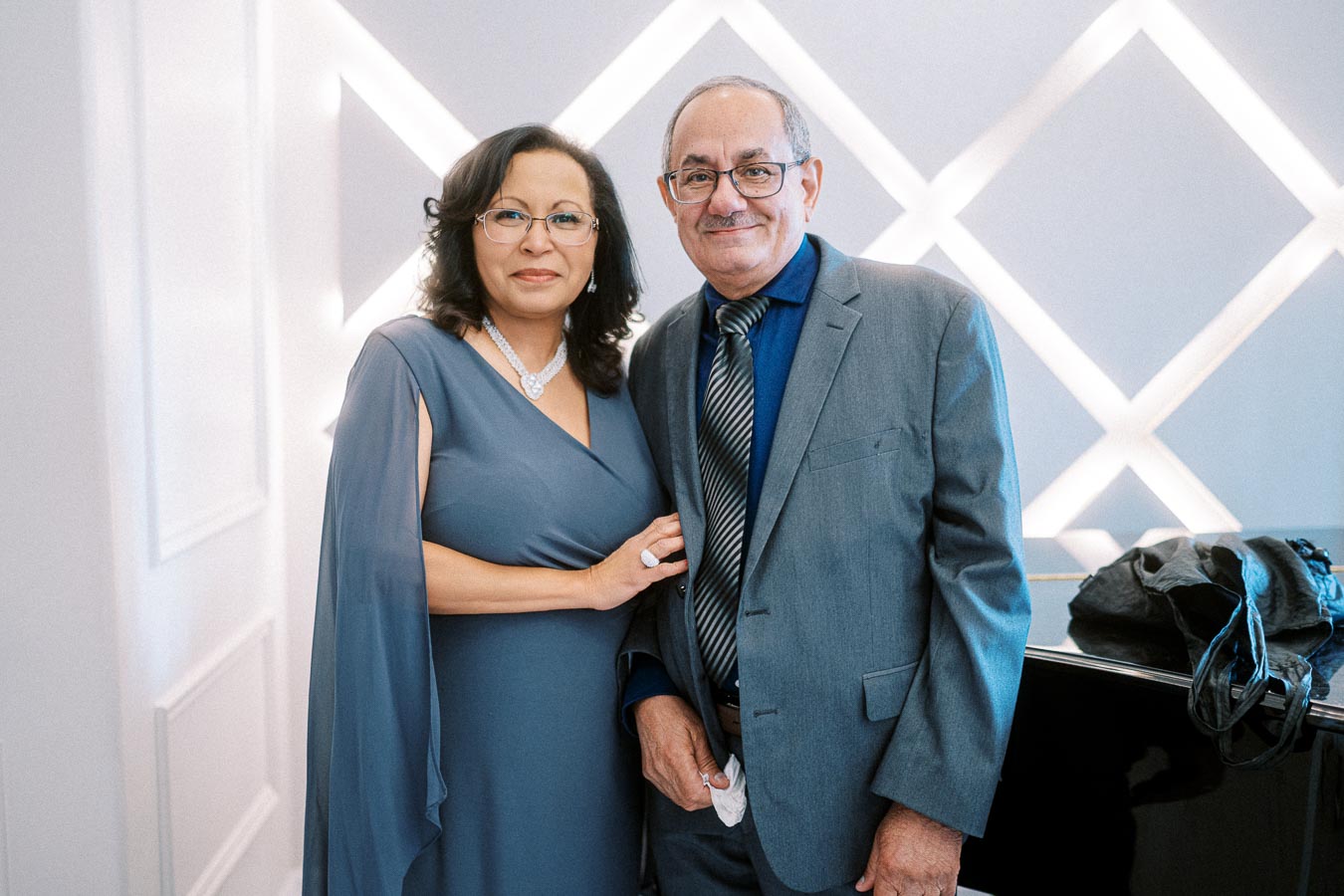 A smiling couple dressed elegantly poses in front of a modern, geometric backdrop. The woman wears a blue gown with a