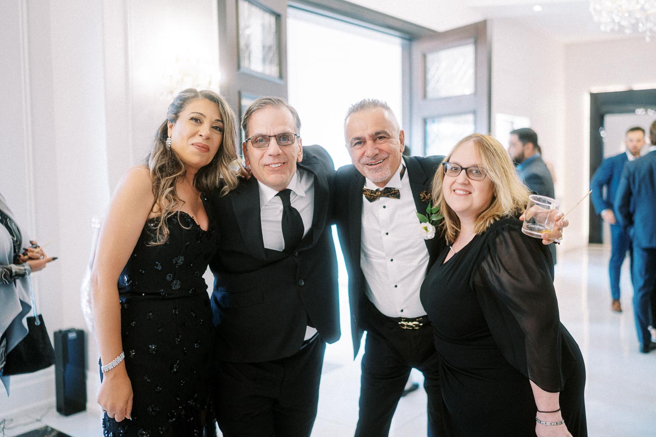 A group of four people cheerfully posing together in formal attire at an indoor event or celebration, with two men in suits