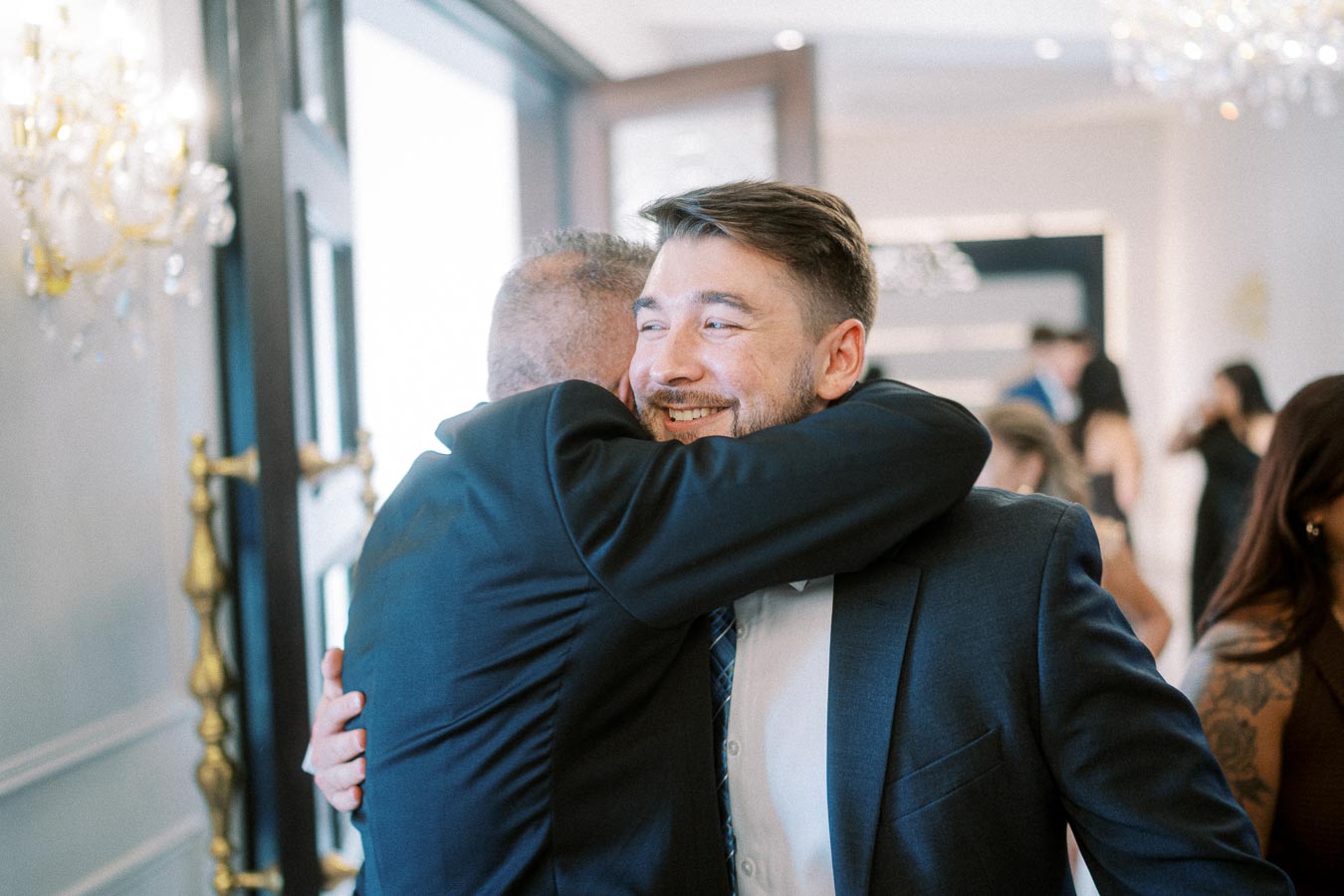 Two men in suits embracing warmly at a social gathering, conveying happiness and camaraderie in a bright, elegant setting.