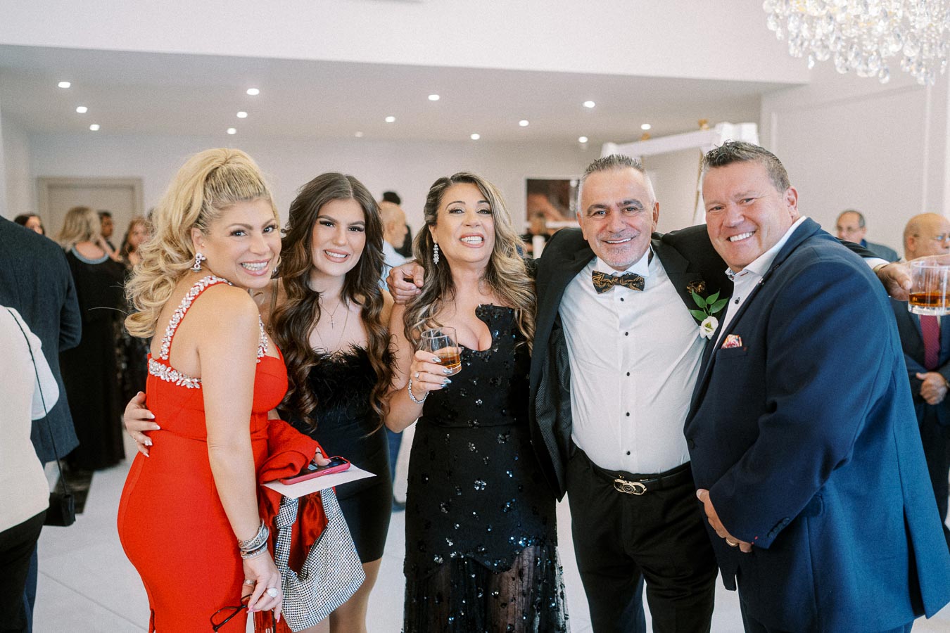 Group of people dressed in formal attire smiling and celebrating at a glamorous indoor event, holding drinks and posing for