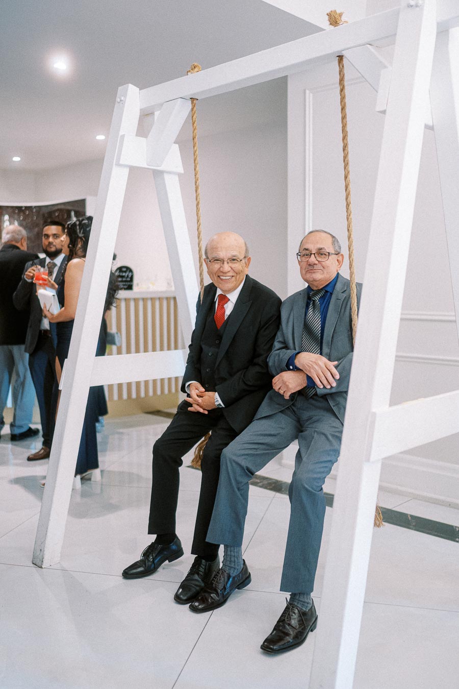 Two elderly men in suits sitting on a large indoor wooden swing, smiling and enjoying a formal event, with other guests in