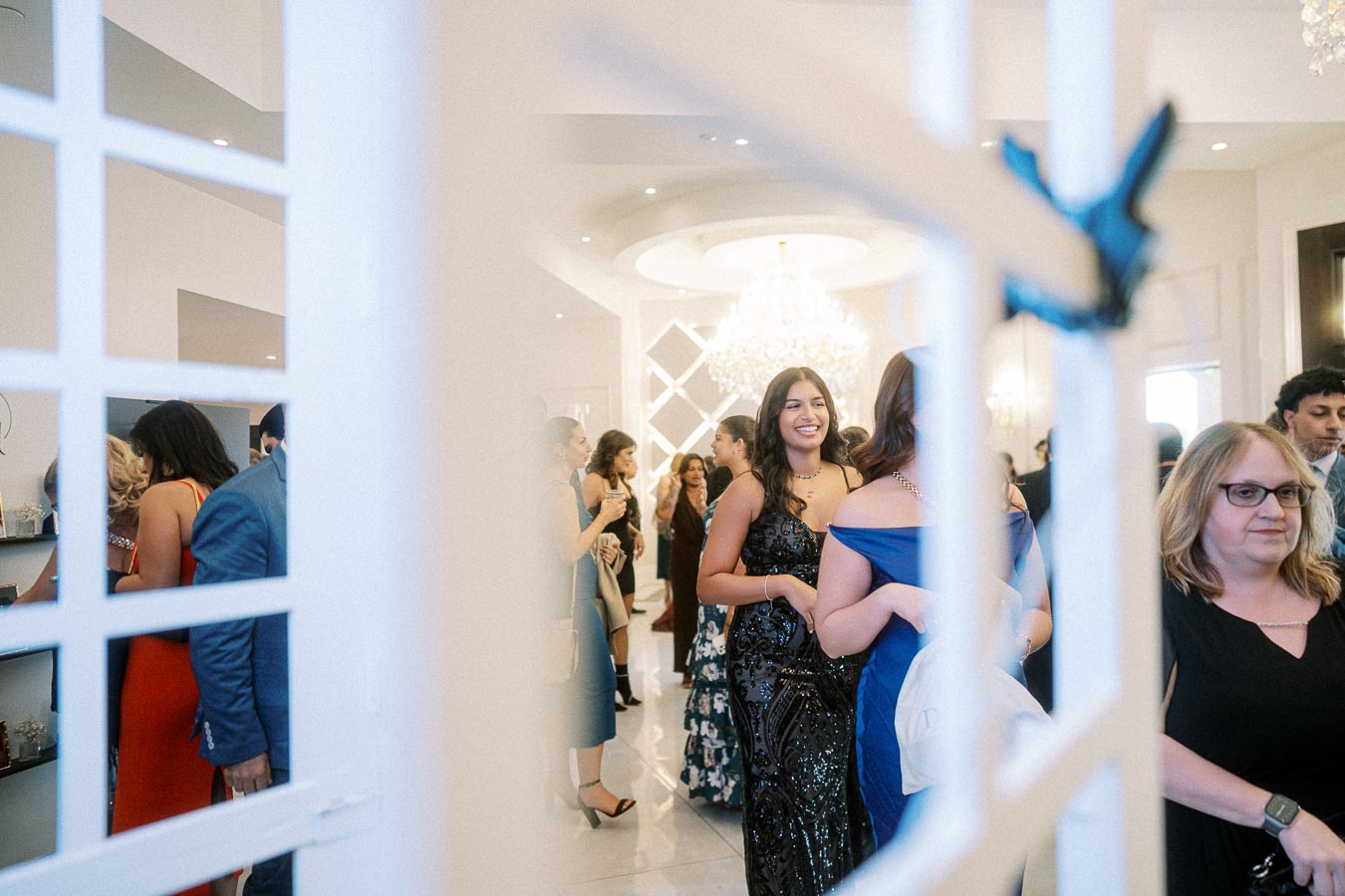 A group of elegantly dressed people socialize at a formal event, with a bright chandelier in the background.