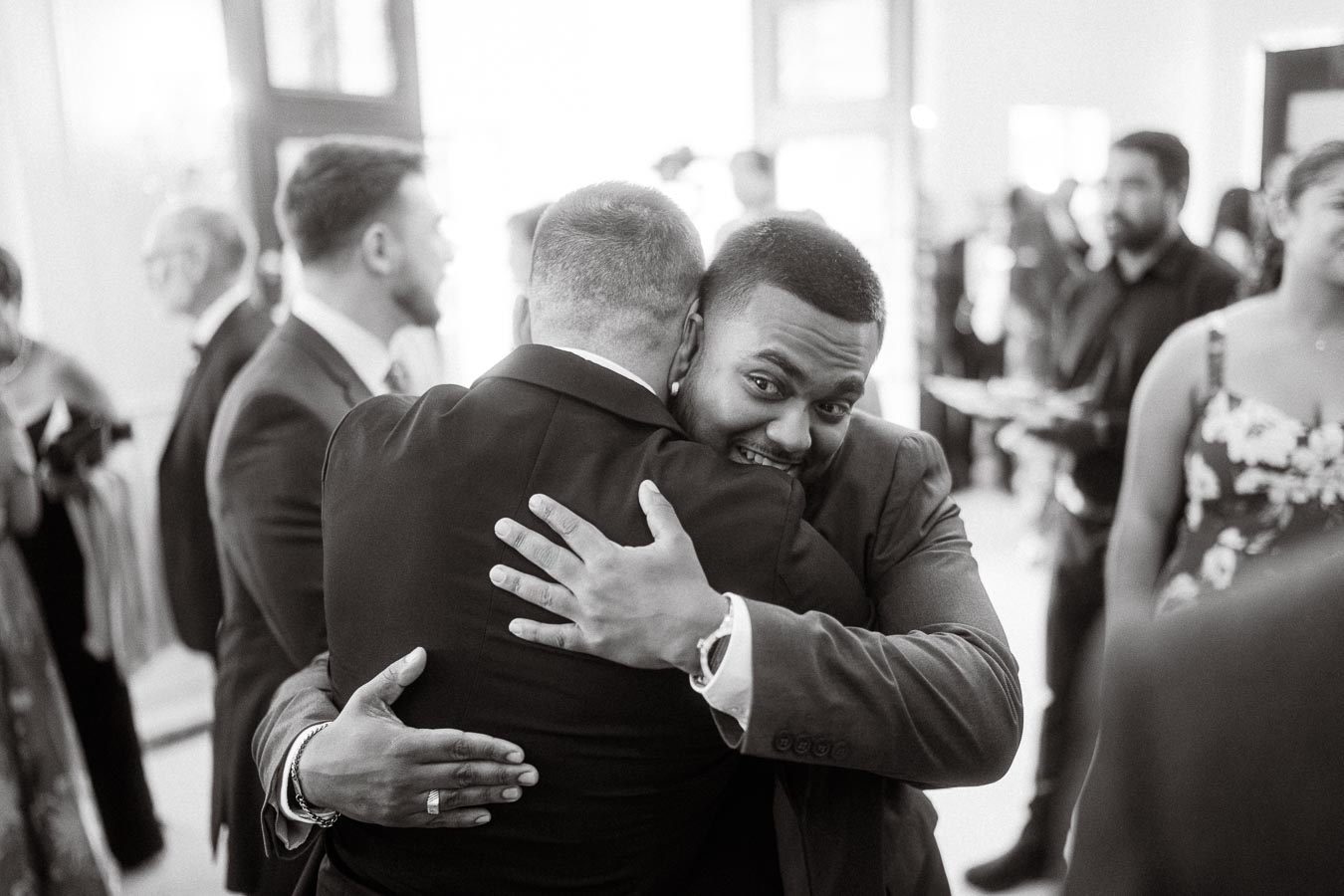 Two men in suits embrace warmly at a formal event, surrounded by other guests dressed in elegant attire. The atmosphere is