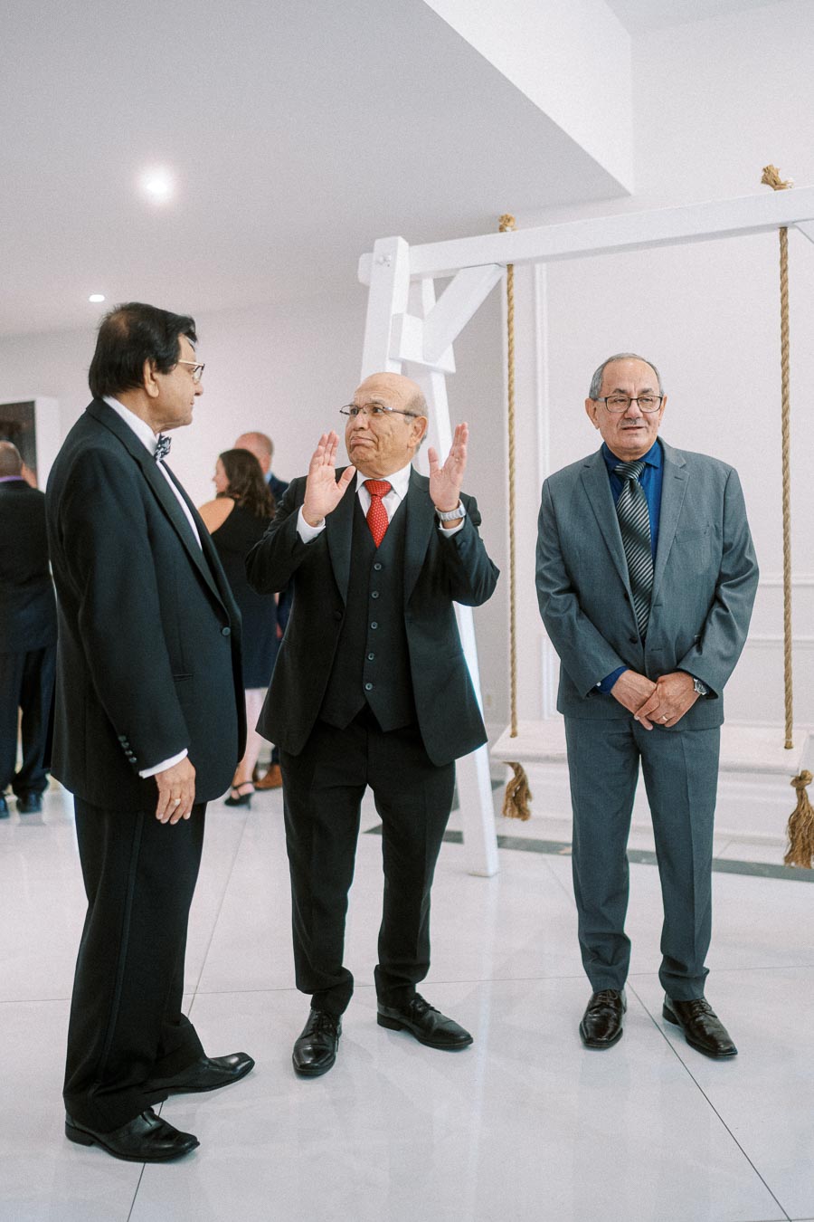 A group of three men in formal attire engaged in conversation at an indoor social event, with a decorative swing in the